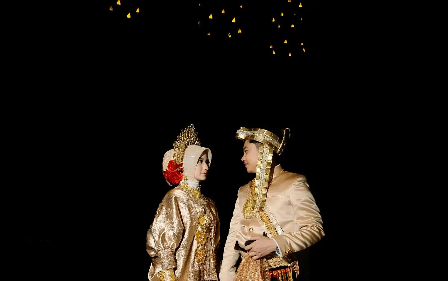Cantika dan Deni di hari pernikahan mereka, berjas bersulam emas dan bersarung tenun merah-emas, saling berhadapan di bawah lampu-lampu kecil keemasan dengan latar hitam