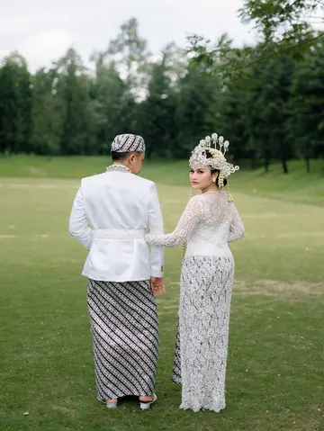 Sepasang pengantin dari belakang di atas rumput, wanita berkebaya renda dan bermahkota, pria berbaju putih dengan sarung batik