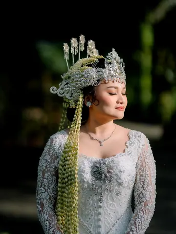 Pengantin wanita bermahkota perak dengan untaian melati panjang dan kebaya renda, mata terpejam, berlatar daun gelap